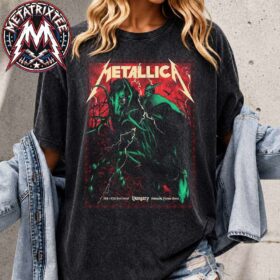 A woman wearing the Metallica M72 Budapest Pop-Up Poster Art By Alex Almeida For June 11th And 13th At Puskas Arena In Hungary Washed T-Shirt with a light blue denim jacket draped over her arms. The heavy metal graphic on the dark, mineral-washed vintage tee captures a glowing green demonic stallion rider under lightning streaks with a red background and the classic bold band typography across the top.